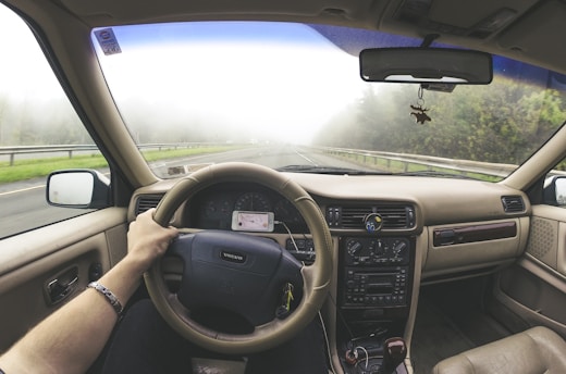 A driver's point of view from inside a car, holding the steering wheel while driving on a foggy road. The car's dashboard displays old-style controls, and a smartphone is mounted nearby. Trees and guardrails line the road outside, creating a serene and slightly mysterious atmosphere.
