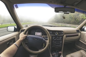 A driver's point of view from inside a car, holding the steering wheel while driving on a foggy road. The car's dashboard displays old-style controls, and a smartphone is mounted nearby. Trees and guardrails line the road outside, creating a serene and slightly mysterious atmosphere.