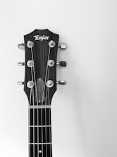 A black and white close-up of a guitar headstock featuring tuning pegs and strings, with a brand name visible on the head. The background is plain and contrasts with the textured wood of the headstock.