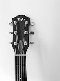 A black and white close-up of a guitar headstock featuring tuning pegs and strings, with a brand name visible on the head. The background is plain and contrasts with the textured wood of the headstock.