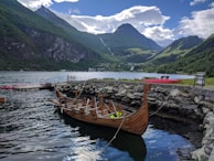 A wooden Viking-style boat is docked beside a stone wall in a tranquil harbor, surrounded by towering, forested mountains. The sky is partly cloudy, with patches of blue peeking through. The water is calm, reflecting the surrounding landscape.