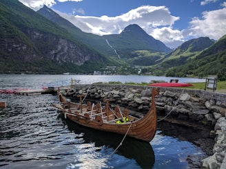 A wooden Viking-style boat is docked beside a stone wall in a tranquil harbor, surrounded by towering, forested mountains. The sky is partly cloudy, with patches of blue peeking through. The water is calm, reflecting the surrounding landscape.