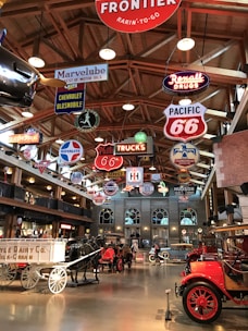 A vintage-themed interior display featuring an array of classic cars, wagons, and bright neon signs hanging from a wooden ceiling. The signs include logos and brand names related to automobiles and the transportation industry. The floor is polished, reflecting the warm light from the fixtures above.