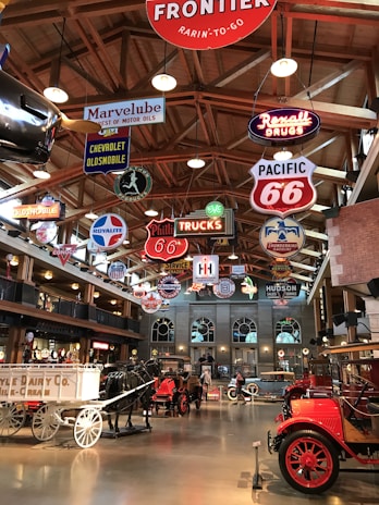 A vintage-themed interior display featuring an array of classic cars, wagons, and bright neon signs hanging from a wooden ceiling. The signs include logos and brand names related to automobiles and the transportation industry. The floor is polished, reflecting the warm light from the fixtures above.