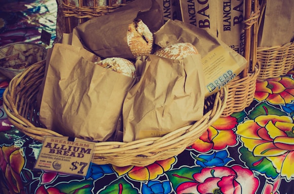 A wicker basket filled with brown paper bags is displayed on a colorful floral tablecloth. Each bag contains a loaf of rustic-looking bread, with some partially peeking out. A paper label attached to the basket reads 'BREADFARM ALLIUM BREAD' and indicates a price. Additional baskets and breads appear in the background.