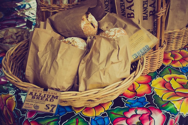 A wicker basket filled with brown paper bags is displayed on a colorful floral tablecloth. Each bag contains a loaf of rustic-looking bread, with some partially peeking out. A paper label attached to the basket reads 'BREADFARM ALLIUM BREAD' and indicates a price. Additional baskets and breads appear in the background.