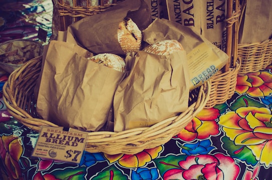 A wicker basket filled with brown paper bags is displayed on a colorful floral tablecloth. Each bag contains a loaf of rustic-looking bread, with some partially peeking out. A paper label attached to the basket reads 'BREADFARM ALLIUM BREAD' and indicates a price. Additional baskets and breads appear in the background.