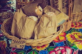 A wicker basket filled with brown paper bags is displayed on a colorful floral tablecloth. Each bag contains a loaf of rustic-looking bread, with some partially peeking out. A paper label attached to the basket reads 'BREADFARM ALLIUM BREAD' and indicates a price. Additional baskets and breads appear in the background.