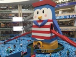 A large penguin-themed inflatable structure in a shopping mall, designed with horizontal red and white stripes and featuring a blue cape. Bright red slides extend from the sides. Surrounding the structure is a pit filled with blue and white plastic balls, with people, including children and adults, interacting with the area. Various stores and advertisements are visible in the background.