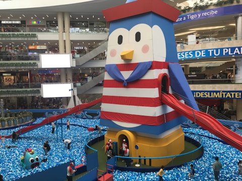 A large penguin-themed inflatable structure in a shopping mall, designed with horizontal red and white stripes and featuring a blue cape. Bright red slides extend from the sides. Surrounding the structure is a pit filled with blue and white plastic balls, with people, including children and adults, interacting with the area. Various stores and advertisements are visible in the background.