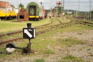 A railway yard with multiple train tracks and various railway equipment. In the foreground, there is a railway signal with the label 'A1a'. A green and red train car is parked in the middle ground. The ground beside the tracks is covered in gravel and grass. In the background, there are more railway carriages, buildings, and a person walking near the tracks.