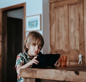 A young child, wearing a floral shirt, is intently looking at a tablet while seated at a wooden table. A plastic cup is also placed on the table. The background includes wooden doors and a framed picture on the wall.