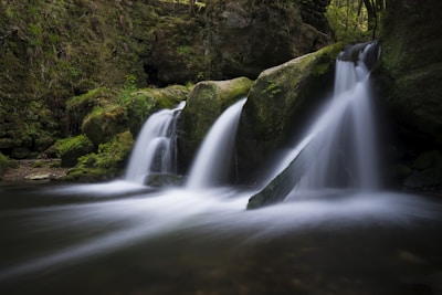 A serene waterfall cascades over moss-covered rocks in a lush, green forest setting. The water flows smoothly, creating a sense of tranquility amidst the vibrant natural environment.
