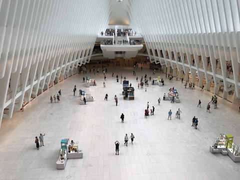 A spacious and modern architectural interior with a high, curved ceiling and a large open floor. Numerous small kiosks are scattered around the area, and several people are walking or standing throughout the space. The design features sleek white structures and columns, creating a futuristic and airy atmosphere.