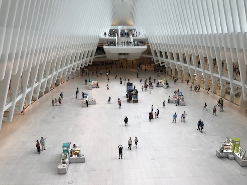 A spacious and modern architectural interior with a high, curved ceiling and a large open floor. Numerous small kiosks are scattered around the area, and several people are walking or standing throughout the space. The design features sleek white structures and columns, creating a futuristic and airy atmosphere.