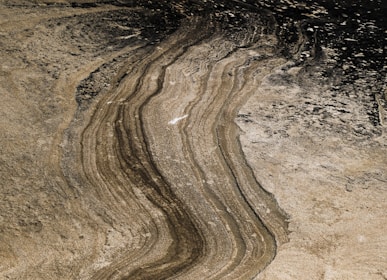 The image features a detailed pattern of natural rock formations with parallel lines and layered textures. Shades of brown, beige, and black give the surface a rugged, weathered look, with visible variations in color due to sedimentation.