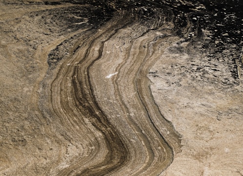 The image features a detailed pattern of natural rock formations with parallel lines and layered textures. Shades of brown, beige, and black give the surface a rugged, weathered look, with visible variations in color due to sedimentation.