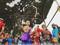 A group of people, including children, are joyfully splashing water and holding colorful bottles under a large canopy. The scene is lively and dynamic, with visible excitement and movement as the liquid sprays outward.