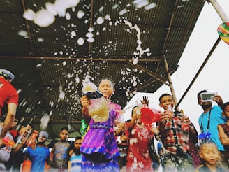 A group of people, including children, are joyfully splashing water and holding colorful bottles under a large canopy. The scene is lively and dynamic, with visible excitement and movement as the liquid sprays outward.