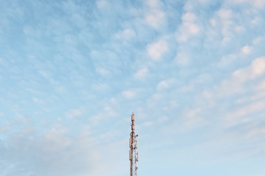 A tall communication tower stands against a backdrop of a blue sky filled with scattered, light clouds. The structure is metallic, with several antennas and equipment visible on its frame.