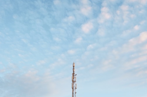 A tall communication tower stands against a backdrop of a blue sky filled with scattered, light clouds. The structure is metallic, with several antennas and equipment visible on its frame.
