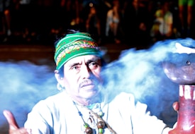 A person wearing a colorful, patterned headband and a white garment, surrounded by a cloud of smoke. The individual appears to be engaged in a traditional or ceremonial activity, holding a vessel emitting the smoke. Bead necklaces add to the cultural attire.