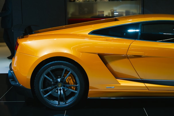 A sleek, high-performance sports car in a vibrant orange color is showcased, highlighting its aerodynamic design and black alloy wheels. The image captures the side view of the vehicle, emphasizing the contours and aggressive styling typical of luxury supercars.