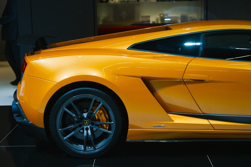 A sleek, high-performance sports car in a vibrant orange color is showcased, highlighting its aerodynamic design and black alloy wheels. The image captures the side view of the vehicle, emphasizing the contours and aggressive styling typical of luxury supercars.