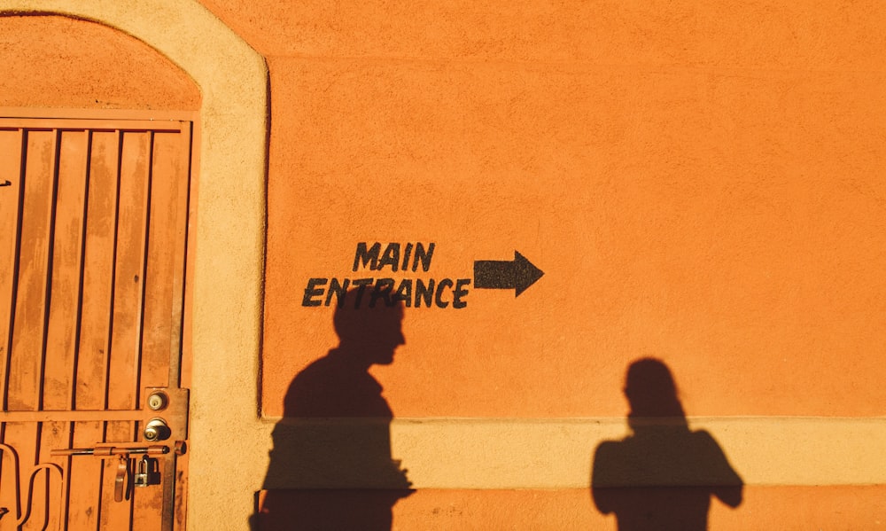 Foto da silhueta de duas pessoas refletindo na parede pintada de laranja com impressão da entrada principal