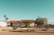 A small house with a red tiled roof, surrounded by a wire fence. The house is set in a dry, desert-like environment with sparse vegetation. There are a few palm trees visible, adding a tropical feel. The sky is clear with a soft blue hue, and there are signs and utility poles visible in the background.