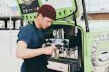 A person wearing a blue shirt and burgundy beanie is preparing coffee using an espresso machine installed in a green mobile cart. They are holding a milk frother and appear focused on making a beverage. The cart has branding and logos related to coffee on the side.