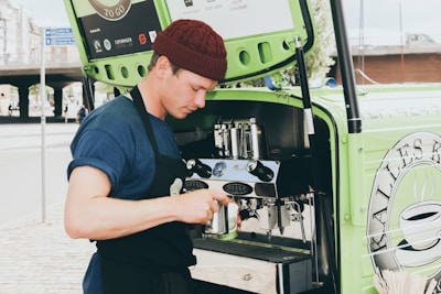 A person wearing a blue shirt and burgundy beanie is preparing coffee using an espresso machine installed in a green mobile cart. They are holding a milk frother and appear focused on making a beverage. The cart has branding and logos related to coffee on the side.