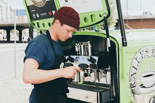 A person wearing a blue shirt and burgundy beanie is preparing coffee using an espresso machine installed in a green mobile cart. They are holding a milk frother and appear focused on making a beverage. The cart has branding and logos related to coffee on the side.