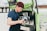 A person wearing a blue shirt and burgundy beanie is preparing coffee using an espresso machine installed in a green mobile cart. They are holding a milk frother and appear focused on making a beverage. The cart has branding and logos related to coffee on the side.