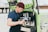 A person wearing a blue shirt and burgundy beanie is preparing coffee using an espresso machine installed in a green mobile cart. They are holding a milk frother and appear focused on making a beverage. The cart has branding and logos related to coffee on the side.