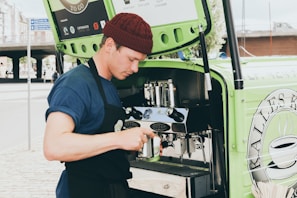 A person wearing a blue shirt and burgundy beanie is preparing coffee using an espresso machine installed in a green mobile cart. They are holding a milk frother and appear focused on making a beverage. The cart has branding and logos related to coffee on the side.