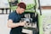 A person wearing a blue shirt and burgundy beanie is preparing coffee using an espresso machine installed in a green mobile cart. They are holding a milk frother and appear focused on making a beverage. The cart has branding and logos related to coffee on the side.