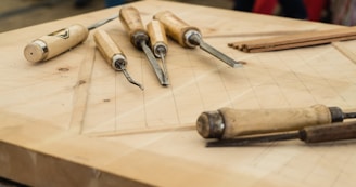 A set of woodworking chisels with wooden handles lies on a flat, wooden surface. The surface is marked with geometric lines, indicating ongoing work on a woodworking project. The handles are light brown, contrasting with the metal blades of the tools. In the background, blurred shapes suggest an indoor workshop setting.