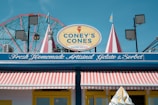 A brightly colored storefront with a sign reading 'Coney's Cones' advertising handmade Italian gelato. The store has a striped awning in red and white, and decorative elements like flags and lights. In the background, a Ferris wheel is visible against a clear blue sky.