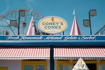 A brightly colored storefront with a sign reading 'Coney's Cones' advertising handmade Italian gelato. The store has a striped awning in red and white, and decorative elements like flags and lights. In the background, a Ferris wheel is visible against a clear blue sky.