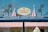 A brightly colored storefront with a sign reading 'Coney's Cones' advertising handmade Italian gelato. The store has a striped awning in red and white, and decorative elements like flags and lights. In the background, a Ferris wheel is visible against a clear blue sky.