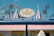 A brightly colored storefront with a sign reading 'Coney's Cones' advertising handmade Italian gelato. The store has a striped awning in red and white, and decorative elements like flags and lights. In the background, a Ferris wheel is visible against a clear blue sky.
