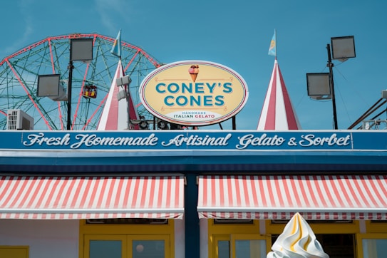 A brightly colored storefront with a sign reading 'Coney's Cones' advertising handmade Italian gelato. The store has a striped awning in red and white, and decorative elements like flags and lights. In the background, a Ferris wheel is visible against a clear blue sky.