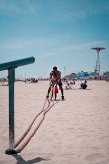 Two people are exercising on a sandy beach by pulling ropes attached to a metal structure. Several beachgoers can be seen in the background. There are umbrellas and other beach accessories scattered around. A tall amusement park structure is visible in the distance.