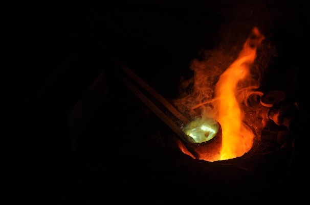 Molten metal is being poured from a crucible under intense heat, creating bright flames and a glowing effect. The scene is dark, with the metal glowing in shades of orange and yellow, highlighting the surrounding area in a dimly lit setting.