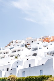 A cluster of whitewashed buildings with flat roofs and arched doorways is set on a hillside under a bright blue sky. The structures reflect a Mediterranean architectural style, typical of a coastal town. Small patches of greenery and vibrant flowers add a touch of color to the predominantly white scene.