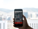 A hand holding a smartphone displaying a map application with a red location pin on the screen. The background features a blurred view of a cityscape with buildings and hills under a cloudy sky.