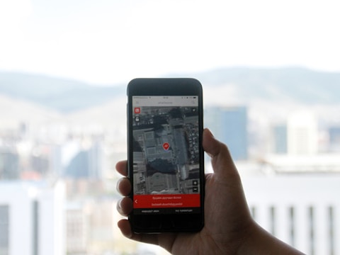 A hand holding a smartphone displaying a map application with a red location pin on the screen. The background features a blurred view of a cityscape with buildings and hills under a cloudy sky.