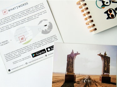 A sheet of paper features information about the application What3Words, including maps and coordinates, with logos for downloading on Google Play and the App Store. Next to it, a colorful notebook with a spiral binding is partially visible. Below these items, there is a photo showing a vast landscape with two ornate stone arches set against an open dirt road leading into the distance under a blue sky.