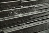 Raindrops splash on a dark metal roof, creating small ripples and droplets. The surface is textured, with water glistening in a dimly lit environment.